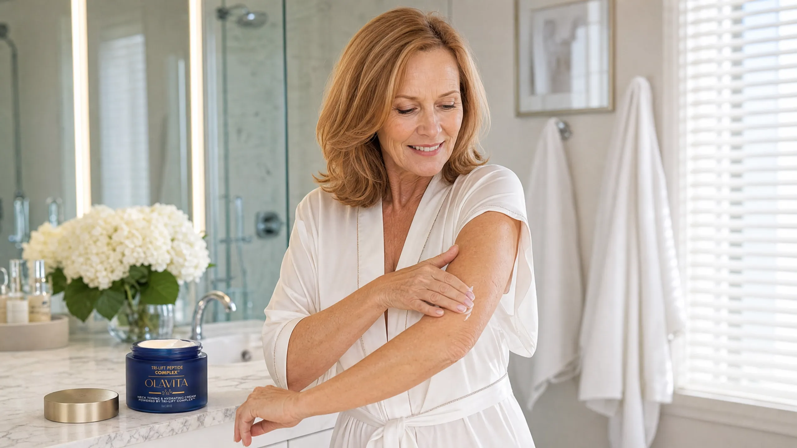 A woman in her sixties applying Olavita cream to her upper arm in a bright bathroom, with the product jar on the counter